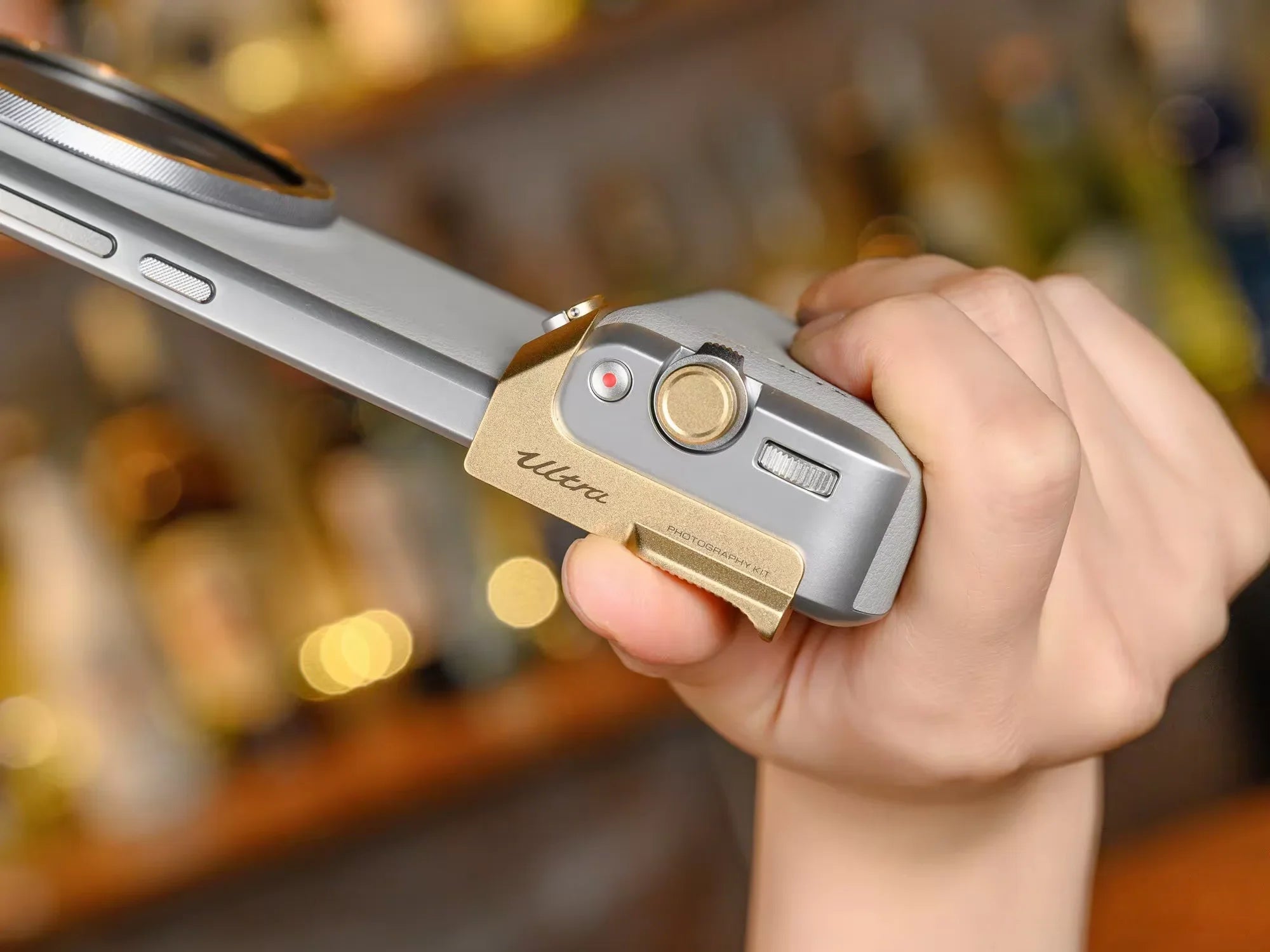Hand holding a silver and gold device with blurred bottles in the background